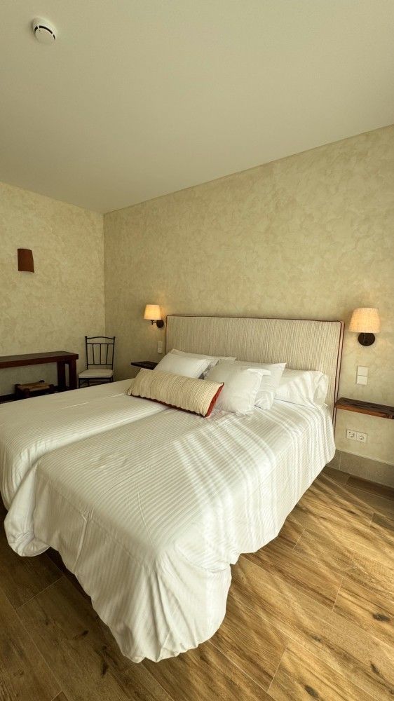 Habitación acogedora con cama blanca, paredes beige, lámparas cálidas y suelo de madera.