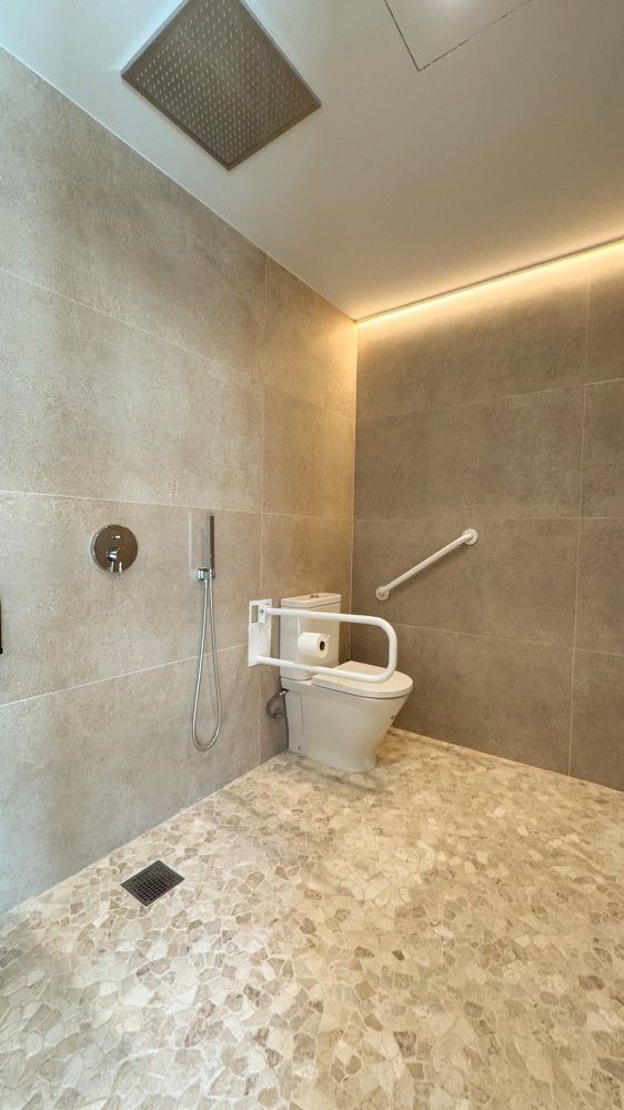Baño accesible con ducha a ras de suelo, inodoro y paredes y suelo de azulejos beige.