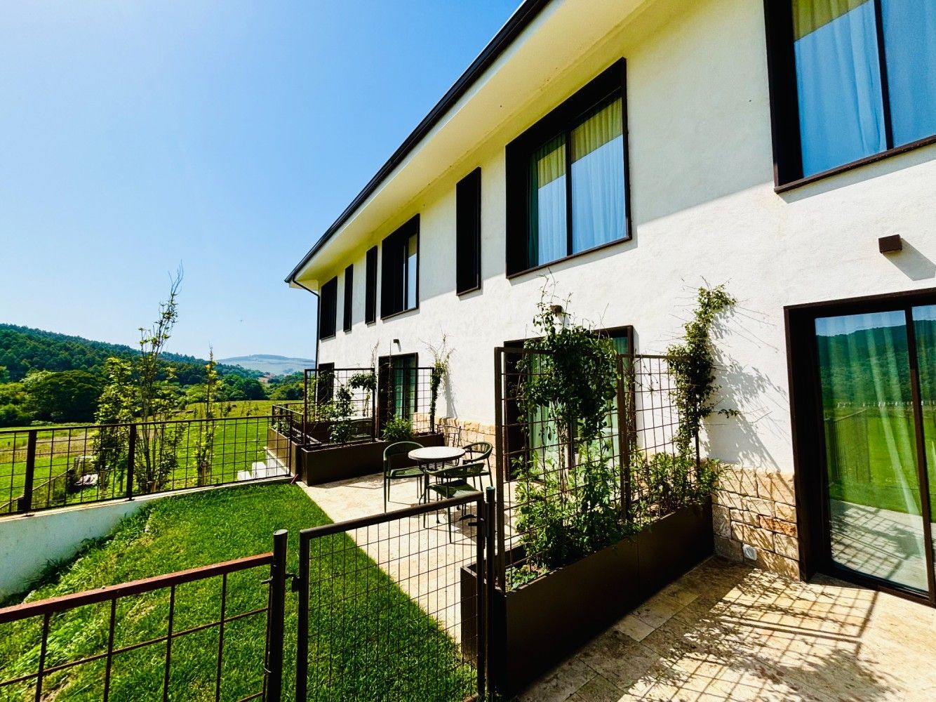 Casa moderna de color blanco con grandes ventanales, terraza y un césped verde con vistas a una ladera soleada.