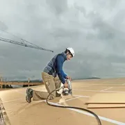 Construction worker kneeling on a rooftop, cutting material with a power saw under a cloudy sky