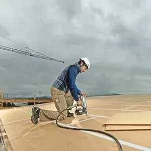 Construction worker kneeling on a rooftop, cutting material with a power saw under a cloudy sky