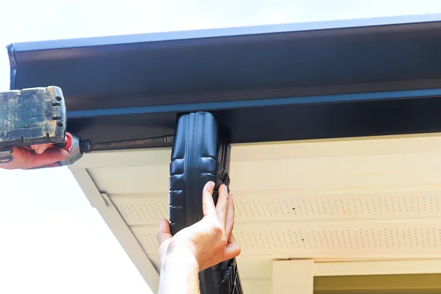 Hands installing a black support bracket under a roof eave on a house
