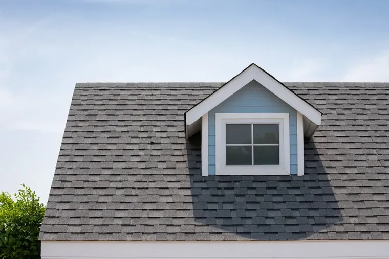 Gray shingled house roof with a small dormer window against a blue sky