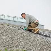 Worker in gray hoodie kneeling on a roof, inspecting shingles with tools nearby
