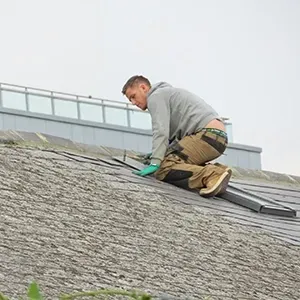 Worker in gray hoodie kneeling on a roof, inspecting shingles with tools nearby