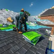 Workers installing green roofing underlayment on a steep roof under a bright blue sky
