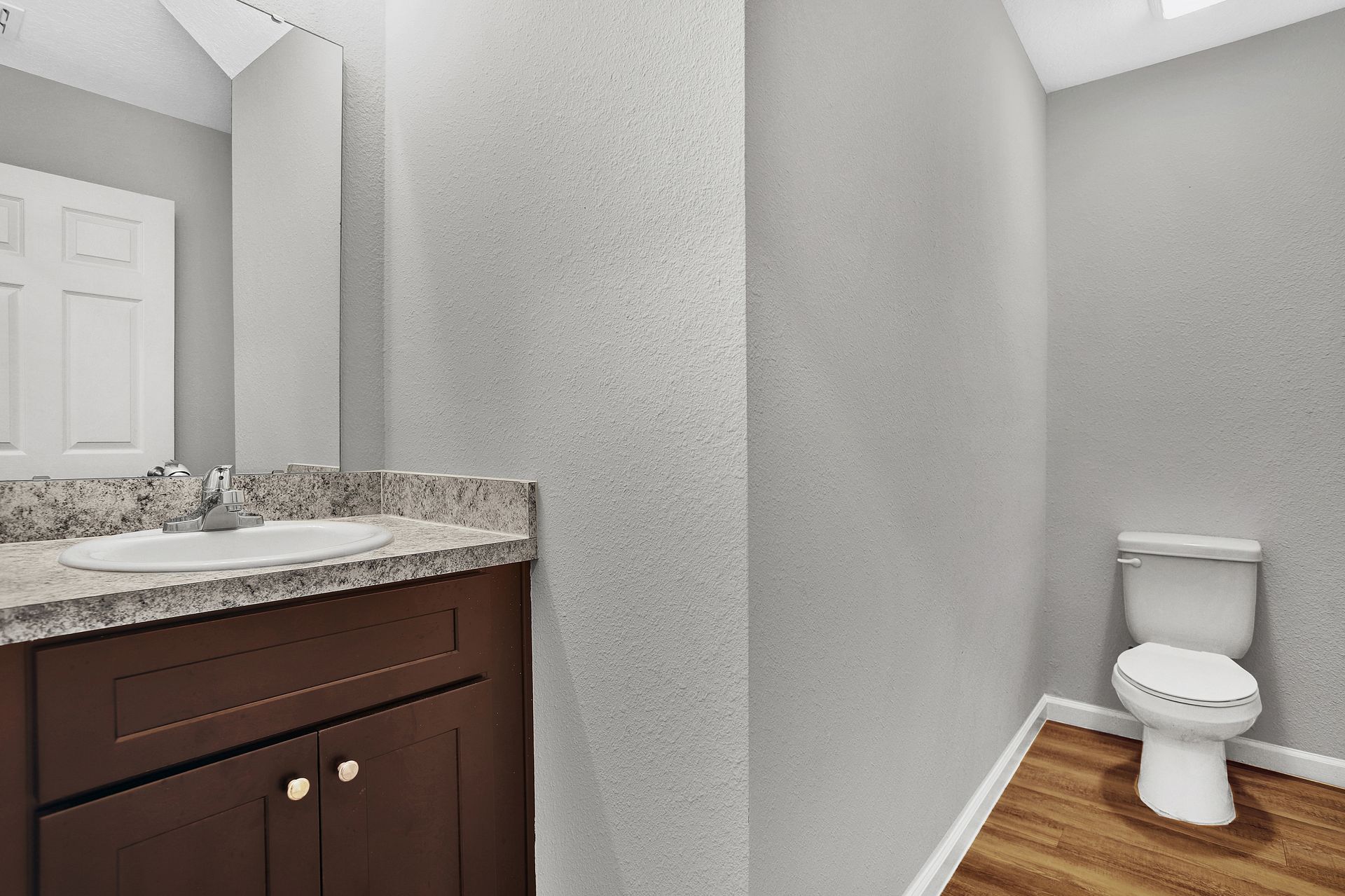 Bathroom with dark brown vanity, granite countertop, white sink, and toilet. Gray walls, wood-look floor.