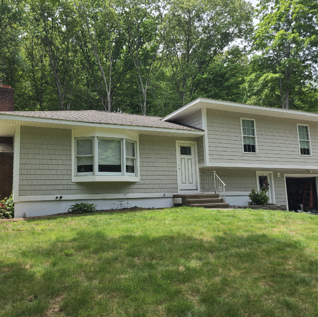 Suburban split-level house with light siding, front lawn, and wooded background