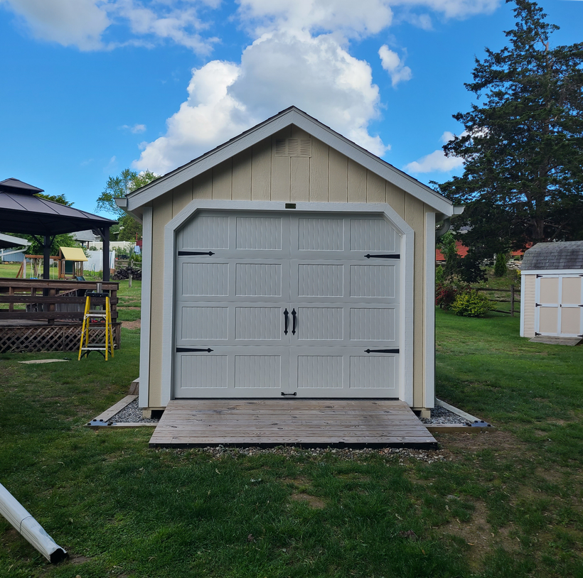 White garden shed with rolling door and a small wooden ramp on a grassy lawn