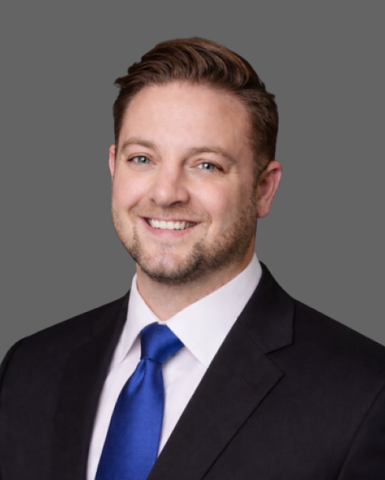 Studio headshot of a smiling professional in a dark suit, white shirt, and blue tie on a gray background