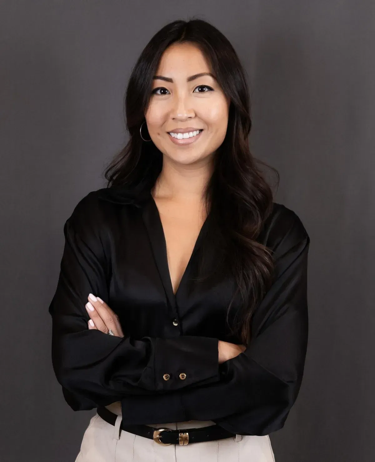Smiling person with arms crossed, wearing a black blouse against a gray background.