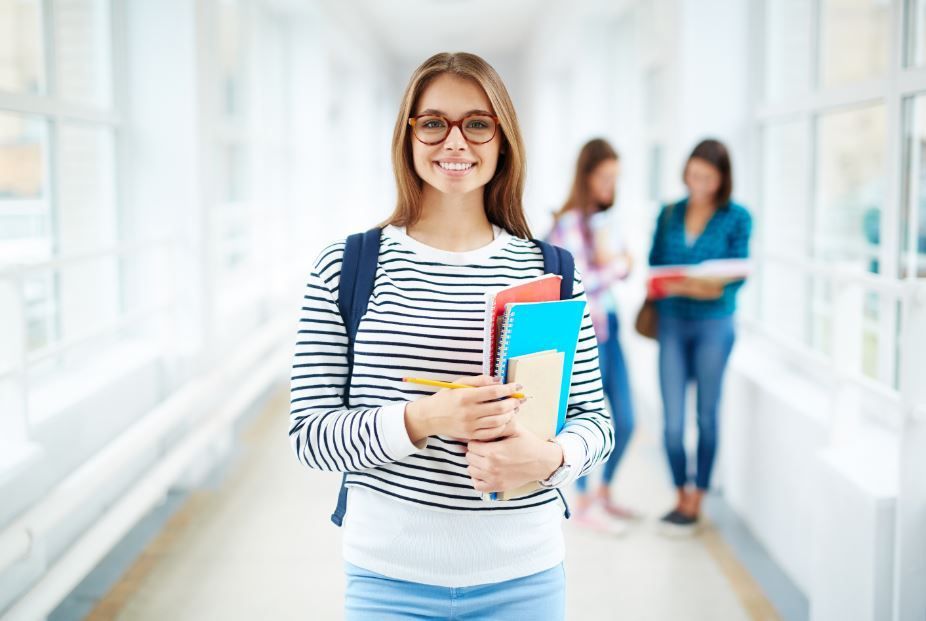 A student smiling and holding books in a bright hallway, with two blurred people talking in the background.