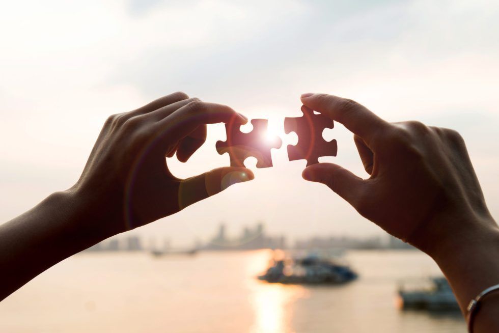 Two hands hold puzzle pieces together against a sunset backdrop over a body of water and a distant city skyline.