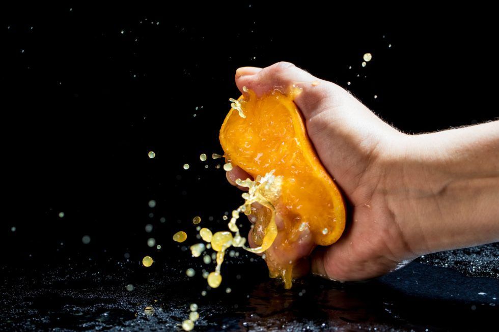 A hand squeezing a fresh orange against a black background, with juice and droplets splashing outward.