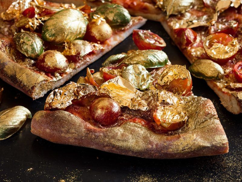 Slices of gourmet pizza topped with roasted vegetables, tomato sauce, and glistening edible gold leaf on a dark surface.