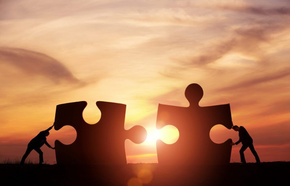 Silhouettes of two people working together to push two large puzzle pieces toward each other against a sunset.