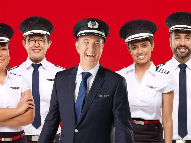 A diverse group of five smiling pilots in white uniforms and peaked caps standing against a solid red background.