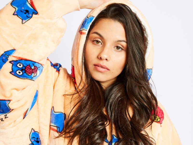 A person with long dark hair wearing a plush, peach-colored hooded sweatshirt patterned with blue and red cartoon cats.