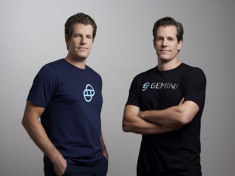 Two men posing against a grey background. One wears a dark blue shirt, the other a black Gemini logo t-shirt.
