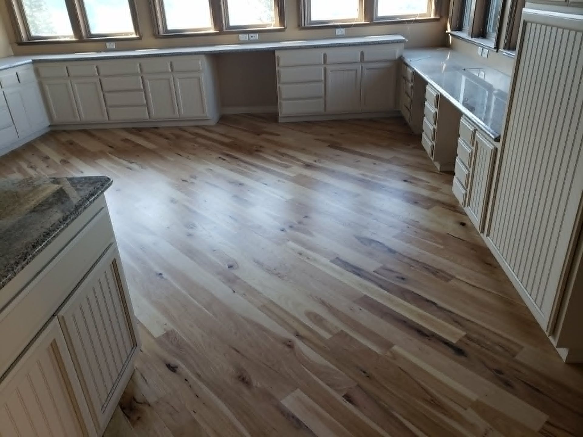 Empty kitchen with light wood floors, white cabinets, and windows along one wall.
