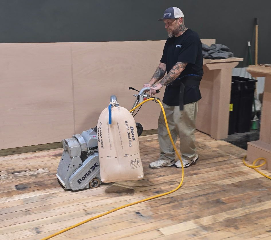 Worker sanding a wooden floor with a floor sander in a workshop, with a hose attached.