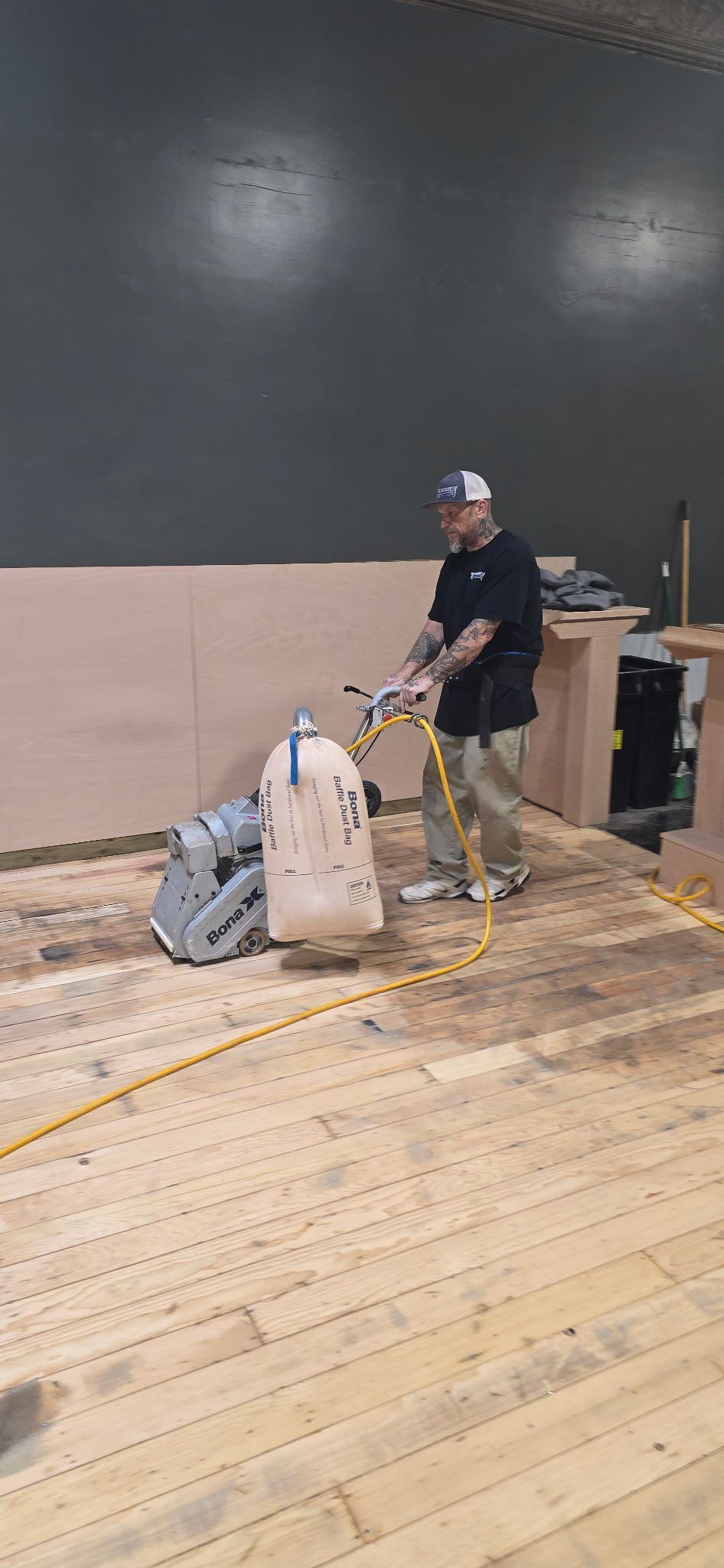 Worker in a room with a gray wall, wooden floor, and supplies stacked beside a cart and trash bin.