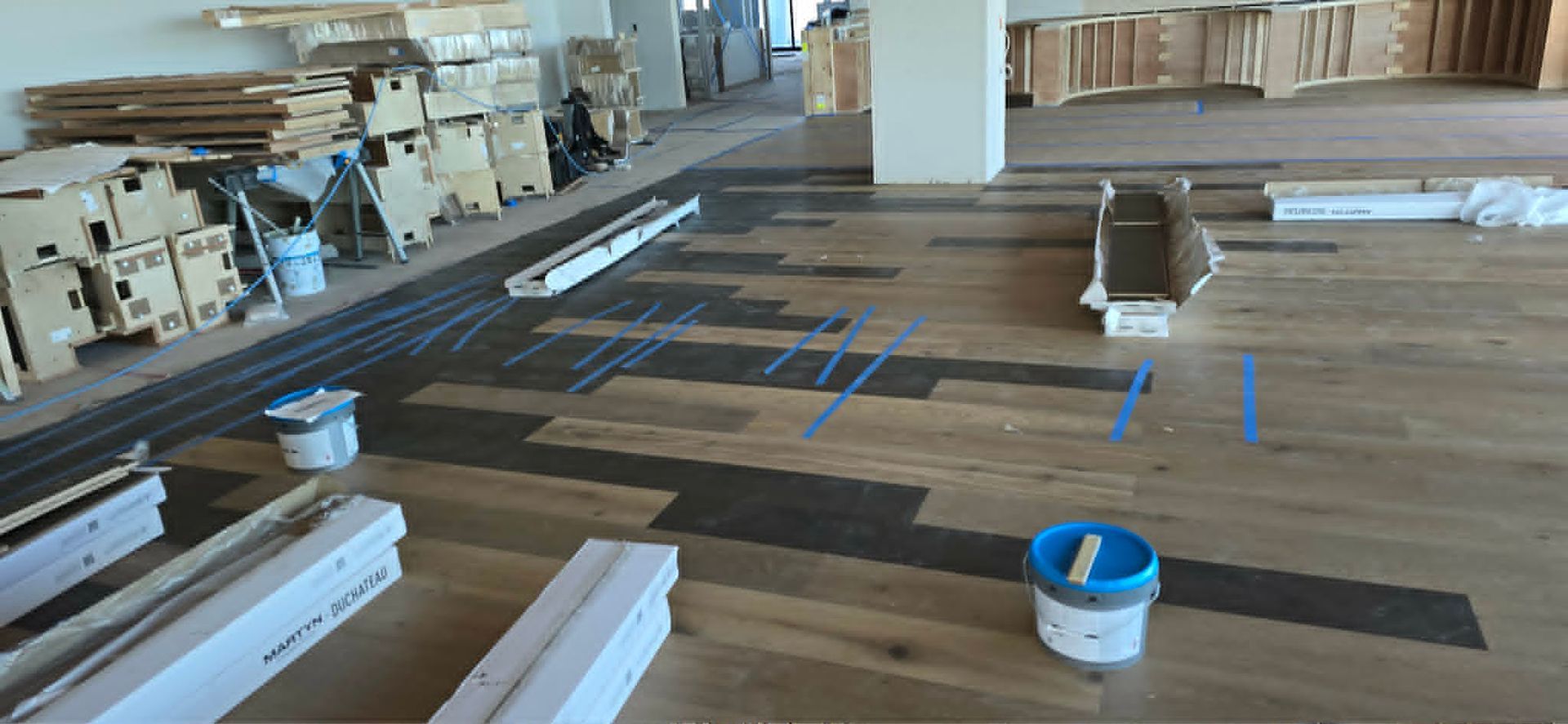 Construction area with wood flooring, white trim pieces, blue tape markings, and a bucket in the foreground