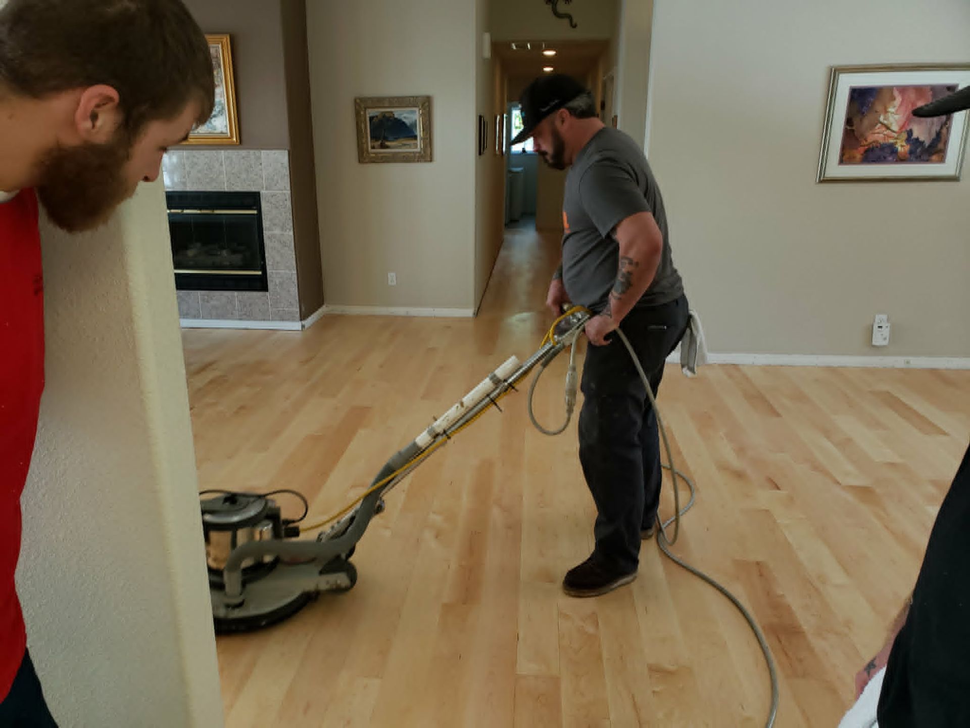 Two men sanding a hardwood floor in an empty room with a floor sander and hose