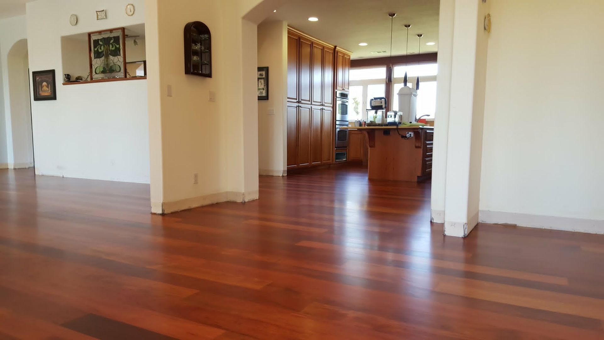 Empty hardwood-floored living room with open doorway to a kitchen and windows in the distance