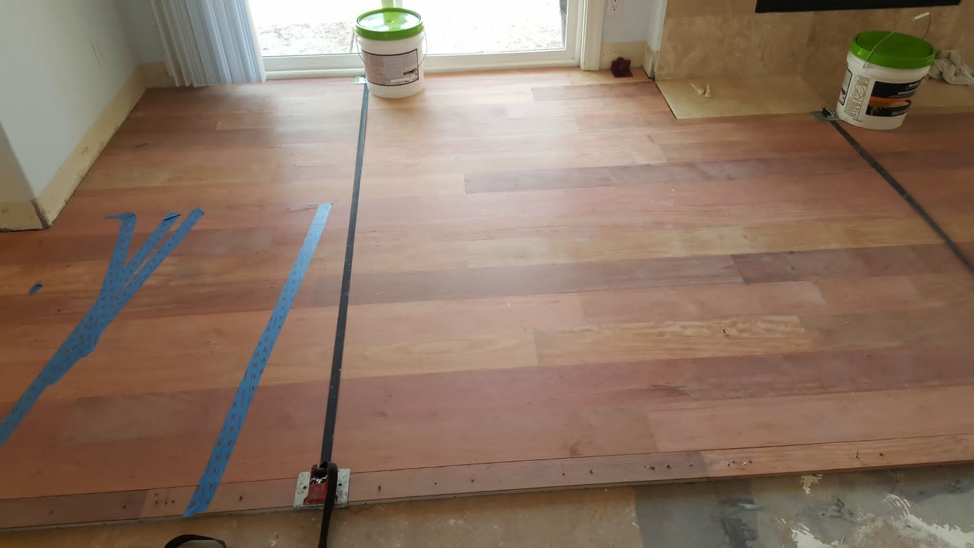 Wooden floor planks being installed in a room, with blue tape markings and a few paint buckets nearby.