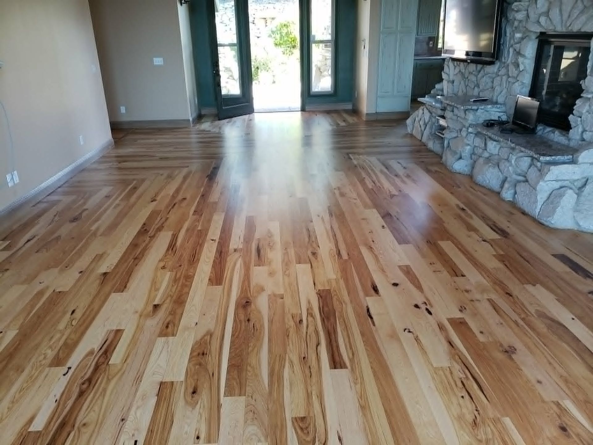 Empty living room with glossy hardwood floors, stone fireplace, and glass doors at the back.