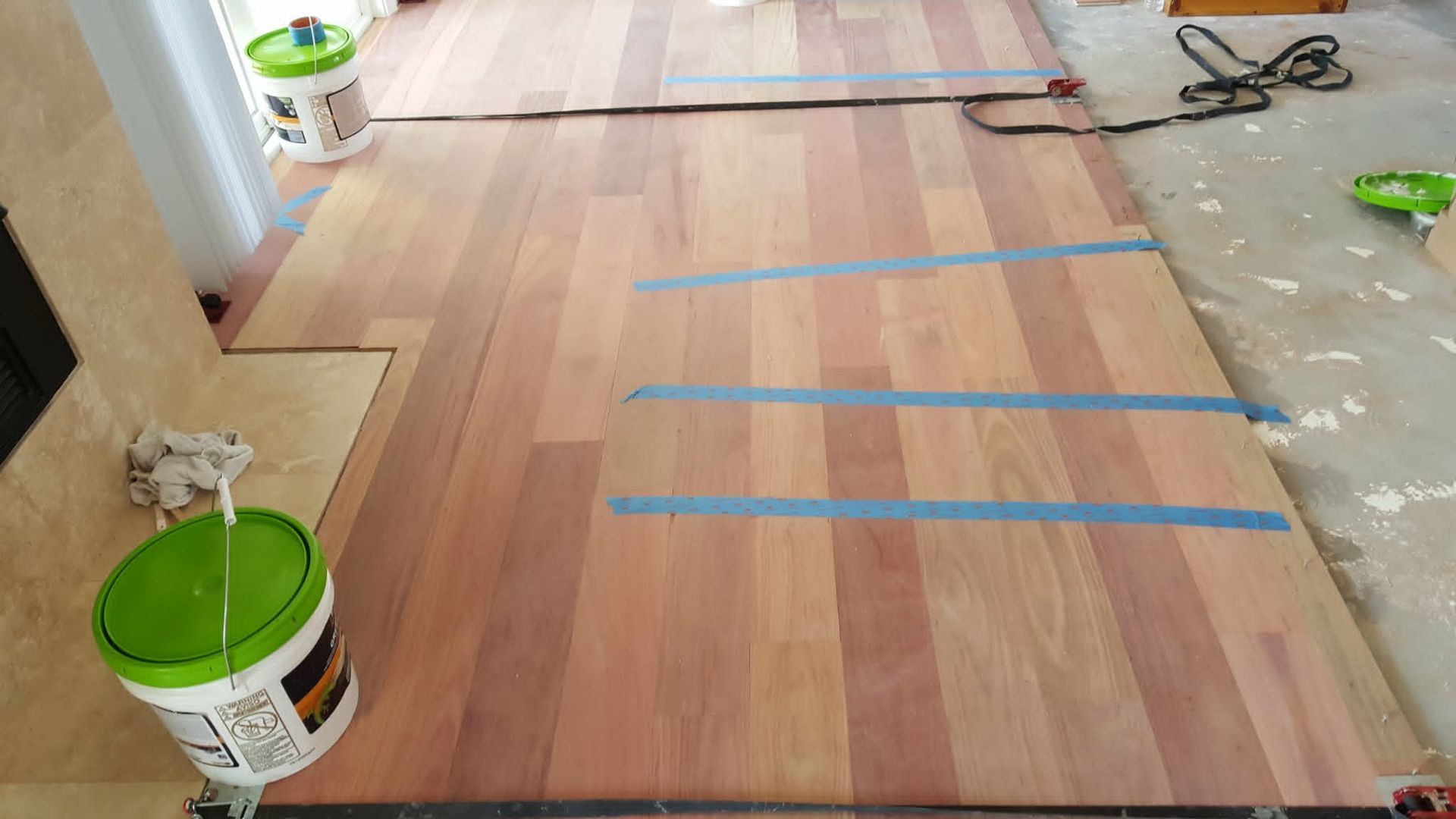 Wood floor under installation with blue tape guides, two paint buckets, and tools near a doorway