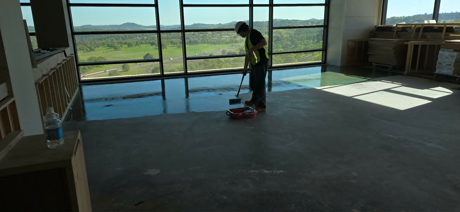 Person sweeping a sunlit room with large windows overlooking a grassy landscape