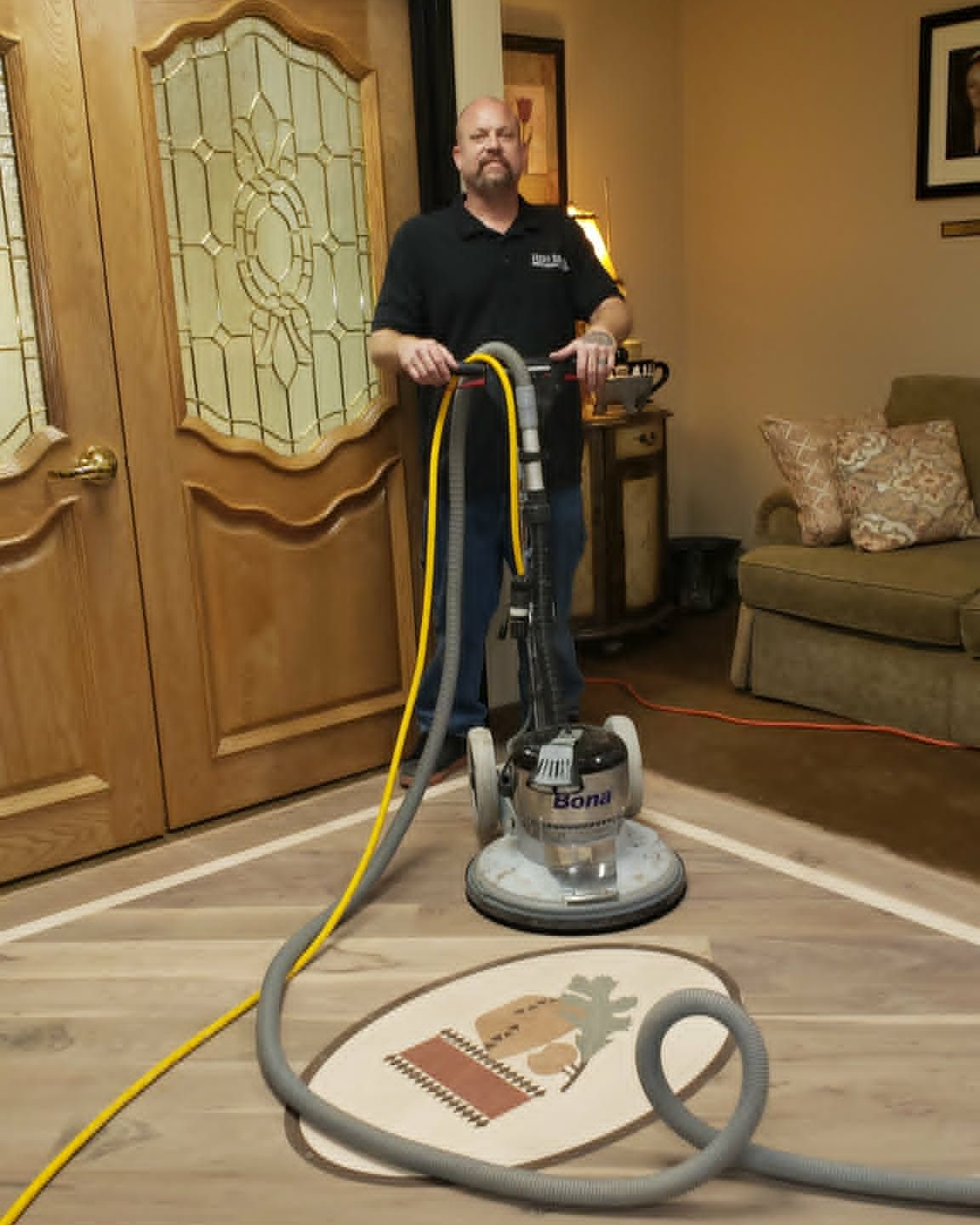 Man standing with a floor sander and hoses in a living room, refinishing hardwood floors