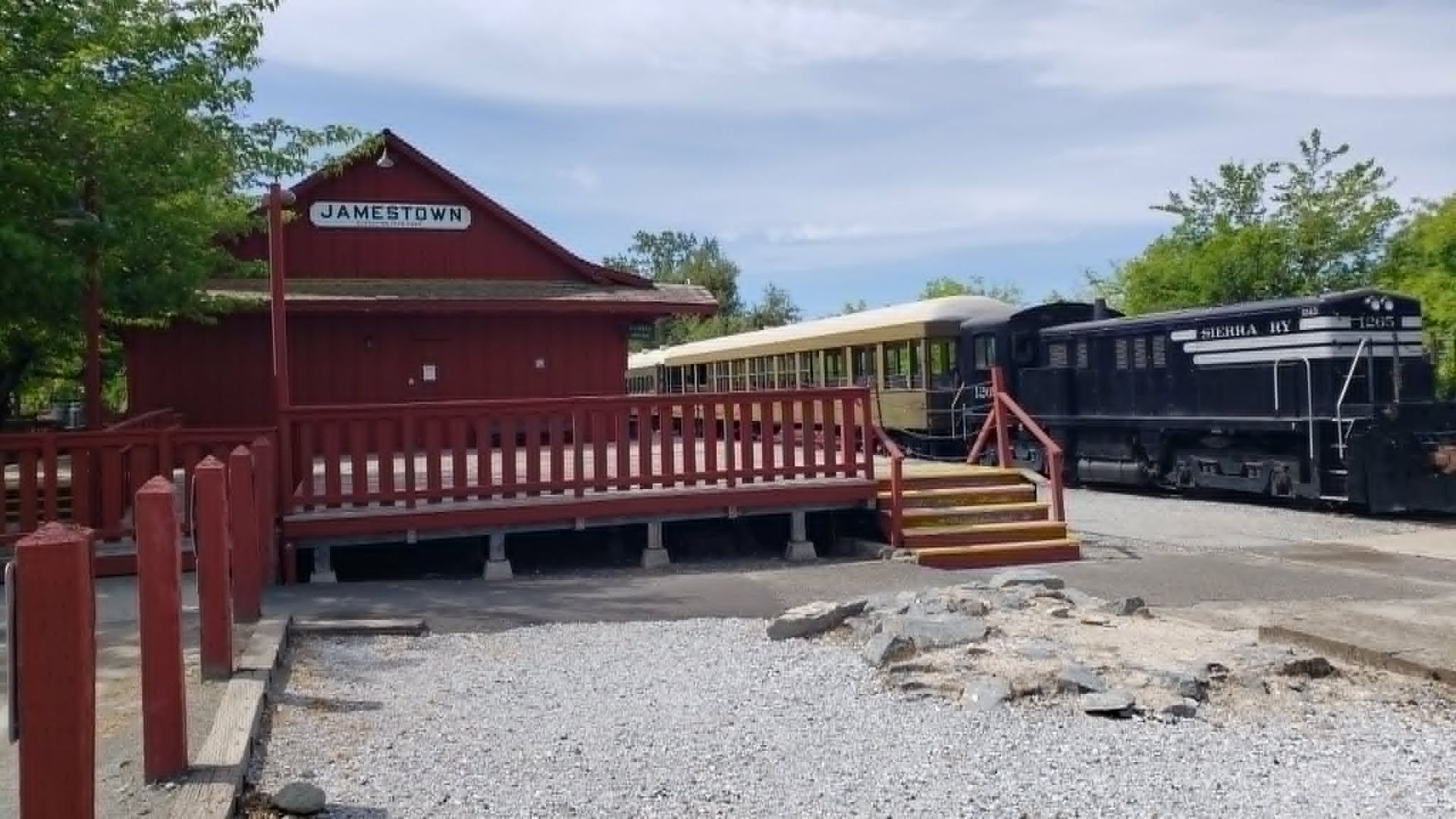 Red wooden depot beside black train cars on a gravel lot under a cloudy sky