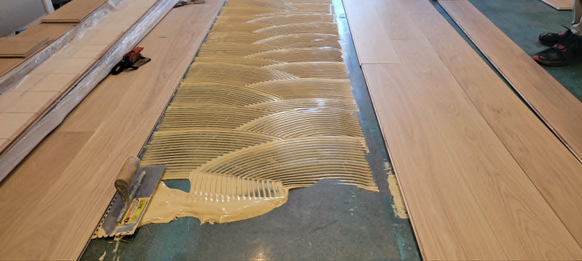 A wood floor strip being glued into place with yellow adhesive along the seam.