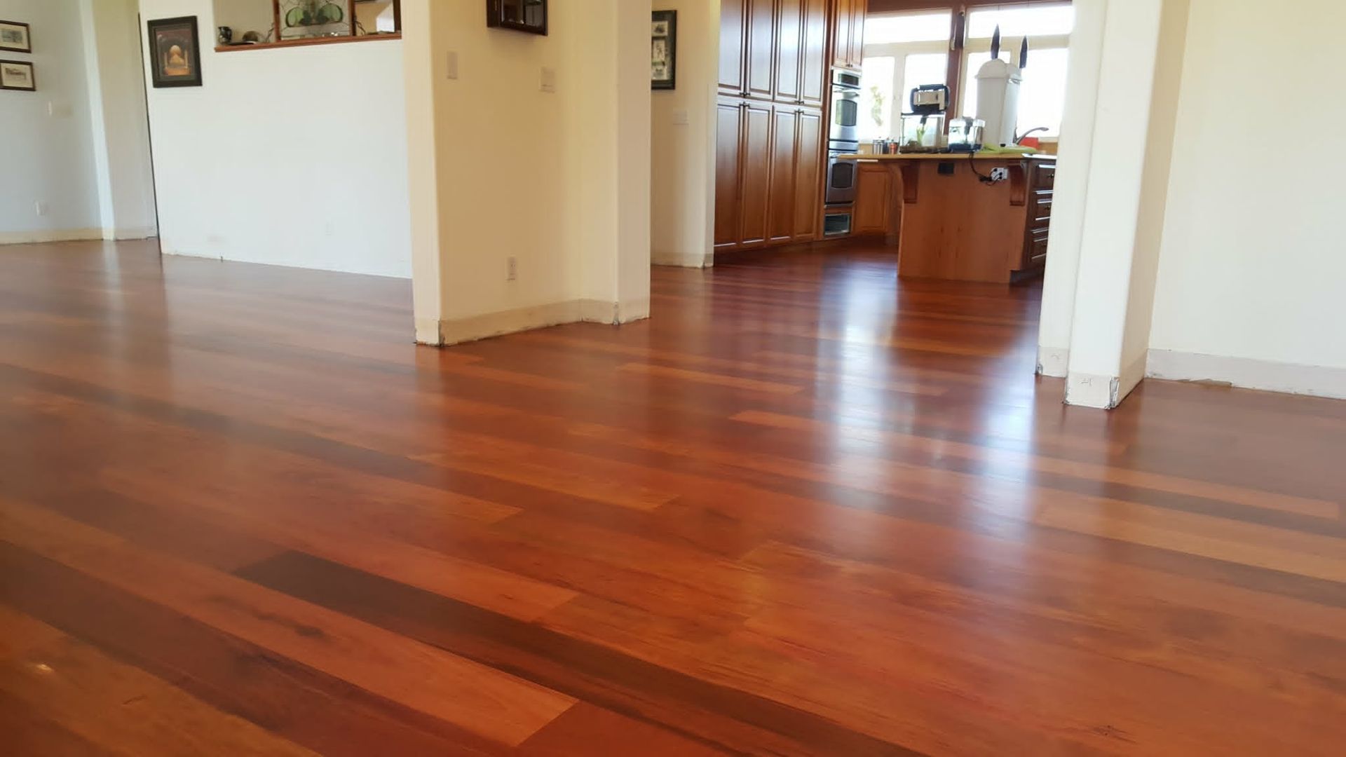 Empty room with polished reddish-brown hardwood floors and an open doorway to a kitchen