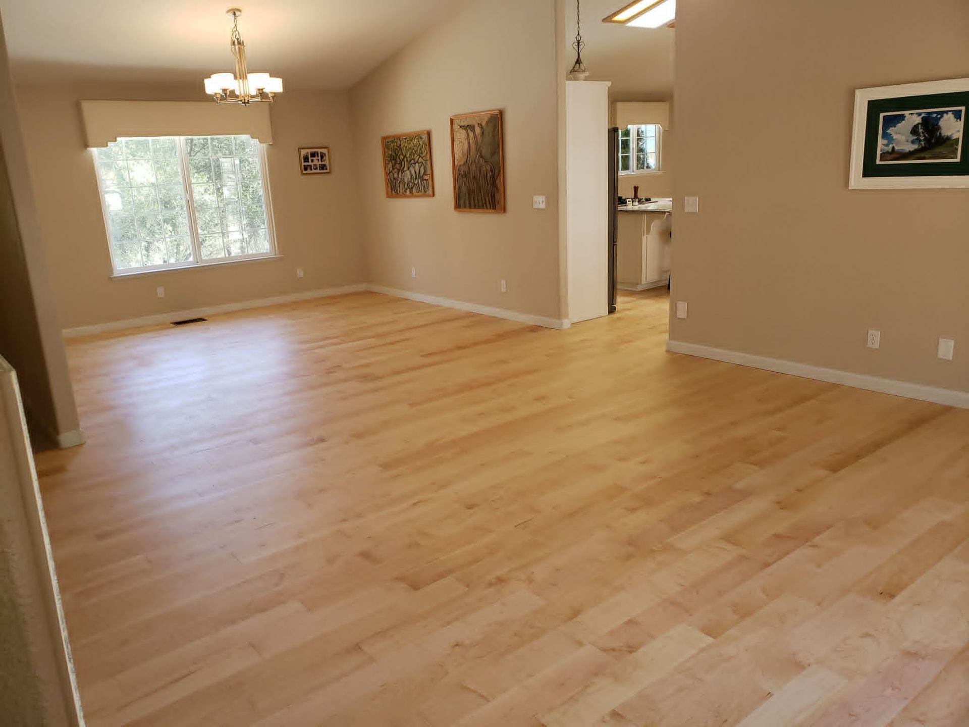 Empty beige dining room with light wood floors, a window, chandelier, and doorway to a kitchen