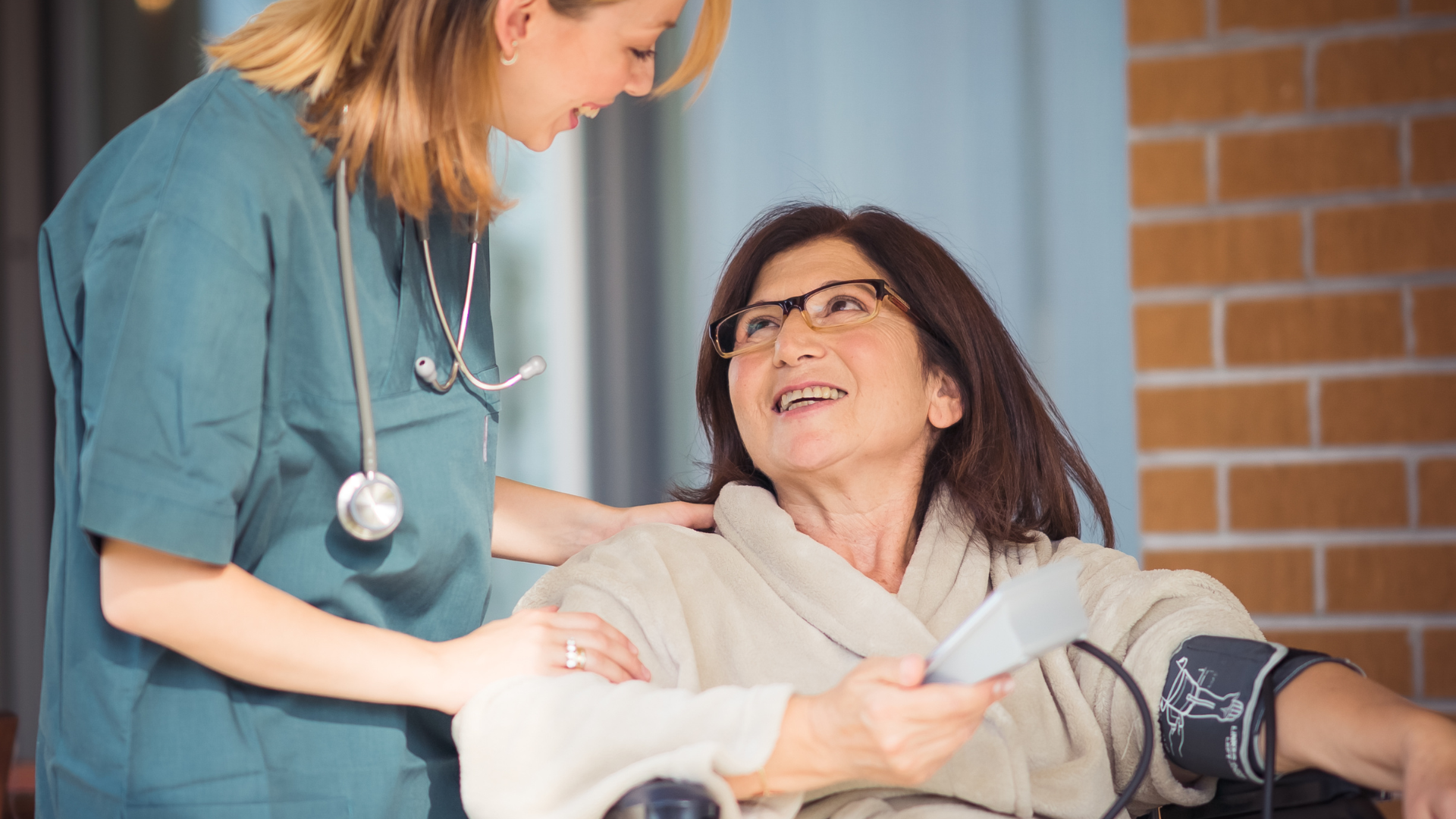 A healthcare worker in teal scrubs talks to a patient wearing a robe, who is having their blood pressure monitored.