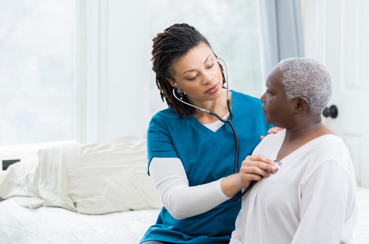 A healthcare professional in a teal scrub top uses a stethoscope to examine an older patient in a white shirt.