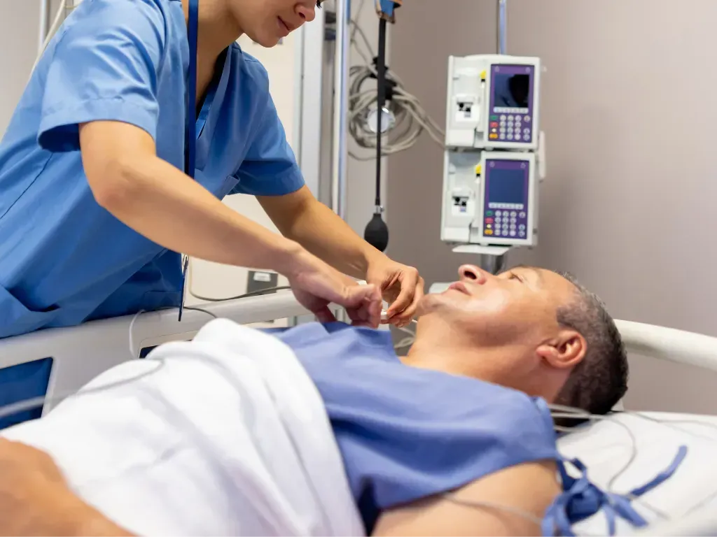 A healthcare professional in blue scrubs attaches medical leads to a patient lying in a hospital bed.