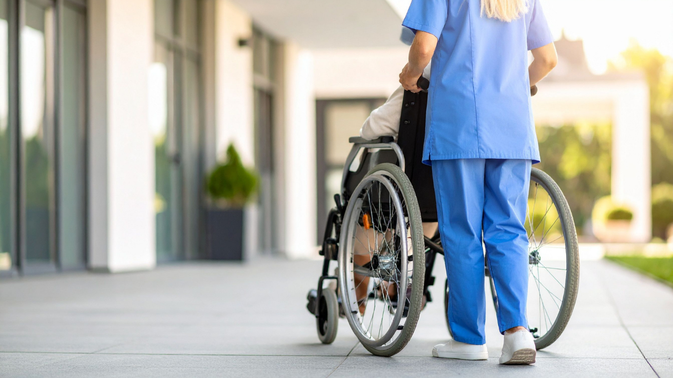 A caregiver in blue scrubs pushes a person sitting in a wheelchair along an outdoor walkway.
