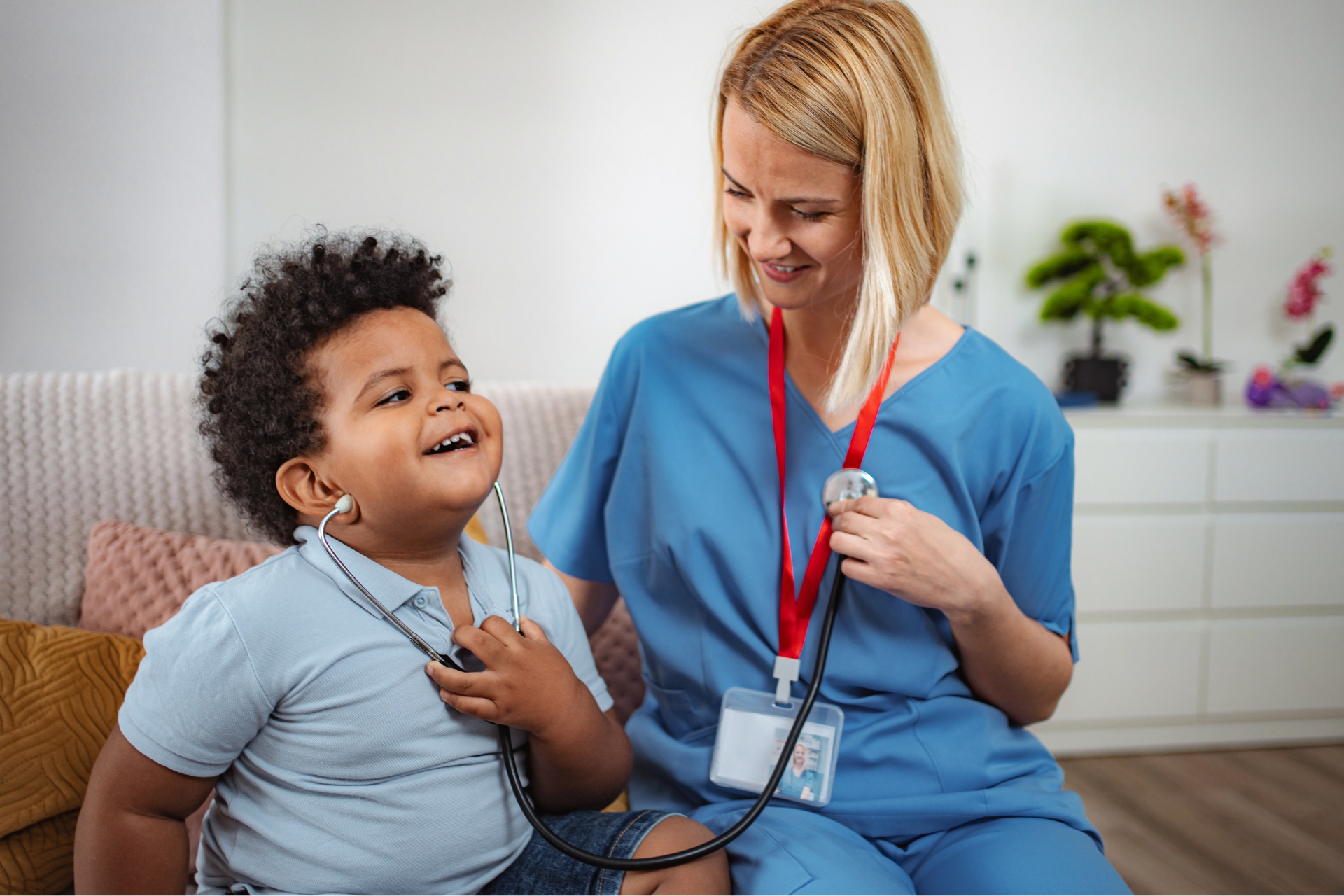 A healthcare worker in blue scrubs smiles as a young child uses a stethoscope to listen to their own heartbeat.