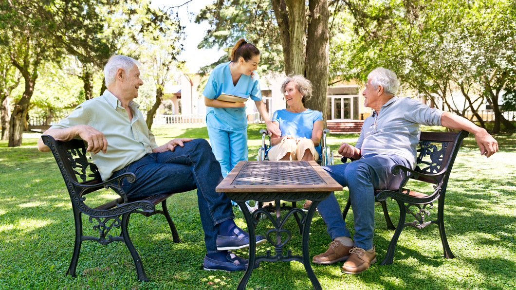 A healthcare worker in blue scrubs interacts with two people sitting on park benches around a small outdoor table.