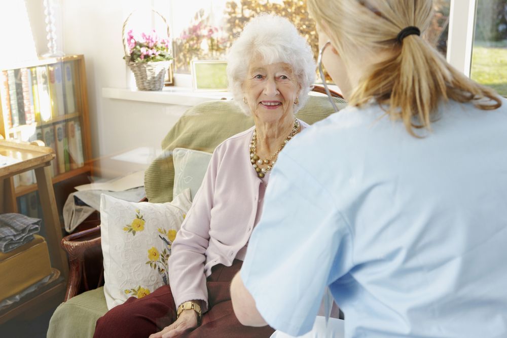 A healthcare professional listens to the heart of an older woman sitting in a chair in a bright, home-like setting.