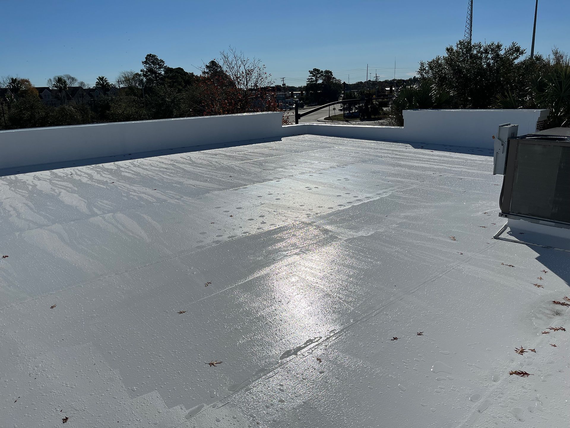 Flat, white rooftop with reflective coating, bright sunlight, surrounding low wall, and trees in background.
