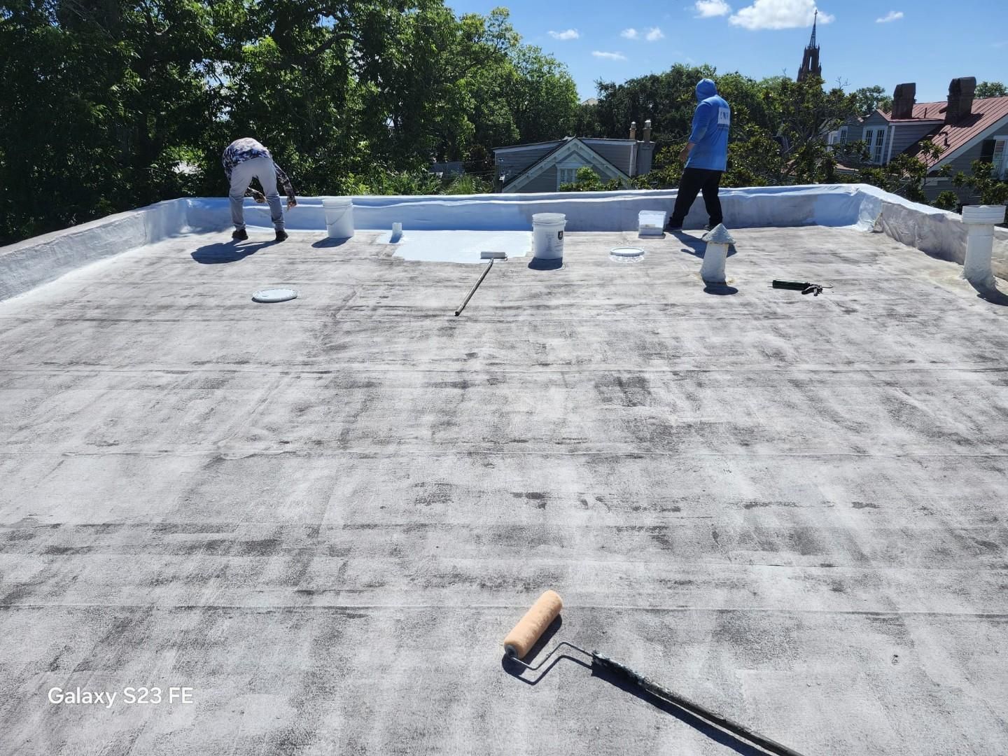 Workers applying white coating to a flat roof. Paint buckets and rollers are visible on the roof under a sunny sky.
