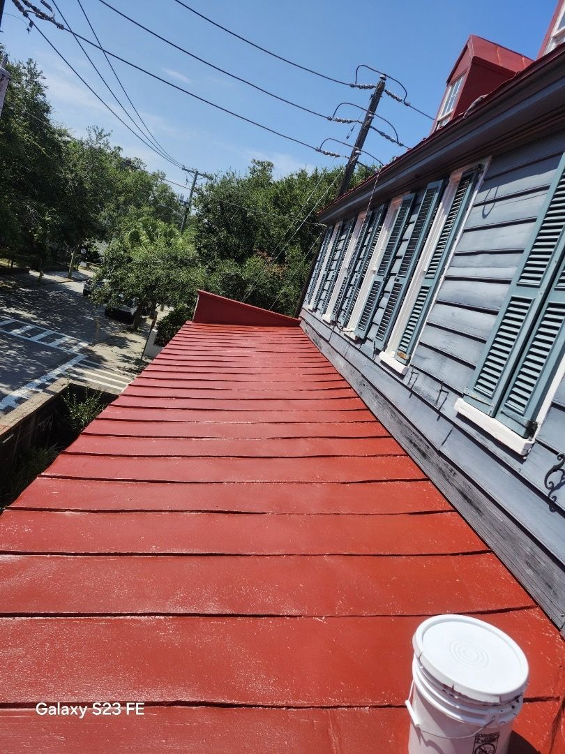 Red metal roof of a house with blue shutters, a white bucket, and power lines against a blue sky.