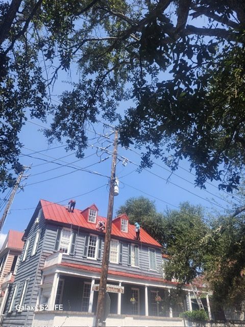 Two-story gray house with red roof, porch, and dormers, under blue sky and tree branches. Utility pole in front.