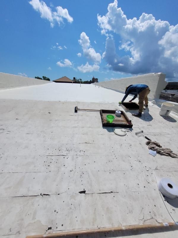 Worker on a white rooftop repairing a roof under a blue sky with clouds.